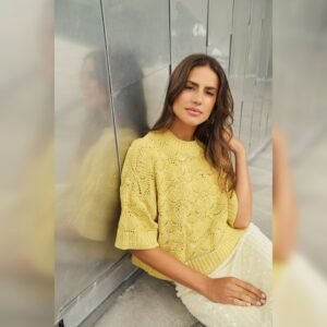 Γυναίκα κάθεται στο πάτωμα δίπλα σε μεταλλικό τοίχο, φορώντας κίτρινο πλεκτό κοντομάνικο μπλουζάκι με ανάγλυφο μοτίβο και λευκή φούστα με παγιέτες. Έχει μακριά καστανά μαλλιά και κοιτάζει με χαλαρή έκφραση προς την κάμερα