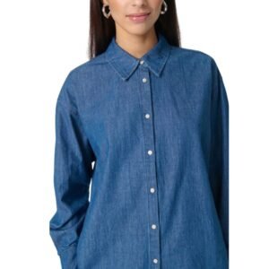 Ημίσωμο πορτραίτο γυναίκας που φορά denim πουκάμισο σε σκούρο indigo χρώμα από 100% βαμβάκι. Το πουκάμισο έχει ίσια εφαρμογή, κουμπιά σε όλο το μήκος και κλασικό γιακά. Η εικόνα έχει καθαρό λευκό φόντο και φυσικό φωτισμό, αναδεικνύοντας την υφή του υφάσματος.