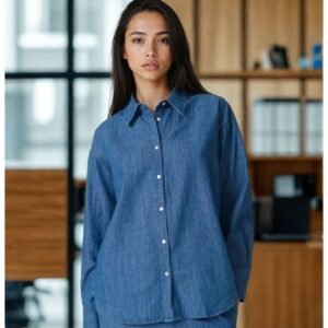 Γυναίκα φορά denim πουκάμισο σε μεσαίο μπλε χρώμα από 100% βαμβάκι. Το πουκάμισο έχει ίσια, χαλαρή εφαρμογή, κλασικό γιακά και κουμπιά σε όλο το μήκος. Το στυλ είναι casual chic, ιδανικό για το γραφείο ή καθημερινές εμφανίσεις. Η φωτογράφιση γίνεται σε εσωτερικό χώρο με φυσικό φωτισμό και φόντο μοντέρνου γραφείου με ξύλινες λεπτομέρειες.