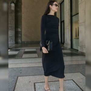 Μαύρο slim fit midi φόρεμα με draped εφαρμογή στη μέση, φορεμένο με minimal σανδάλια και μαύρη τσάντα, σε εξωτερικό σκηνικό με πέτρινους τοίχους