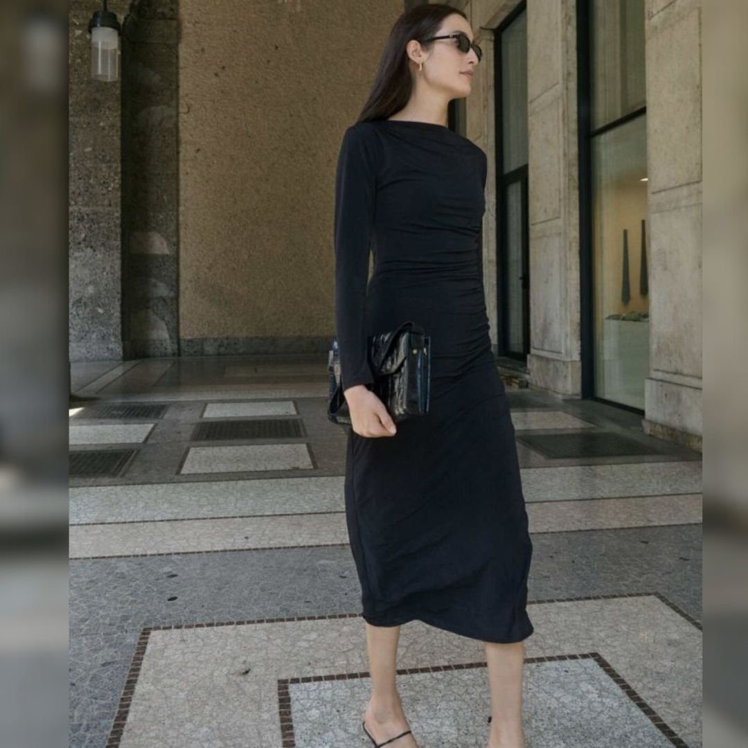 Μαύρο slim fit midi φόρεμα με draped εφαρμογή στη μέση, φορεμένο με minimal σανδάλια και μαύρη τσάντα, σε εξωτερικό σκηνικό με πέτρινους τοίχους