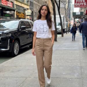 Γυναίκα περπατά σε αστικό πεζοδρόμιο φορώντας λευκό T-shirt με minimal γεωμετρικό σχέδιο και μπεζ παντελόνι σε ίσια γραμμή, συνδυασμένο με λευκά sneakers. Αστικό σκηνικό πόλης με κτίρια και καταστήματα στο βάθος.