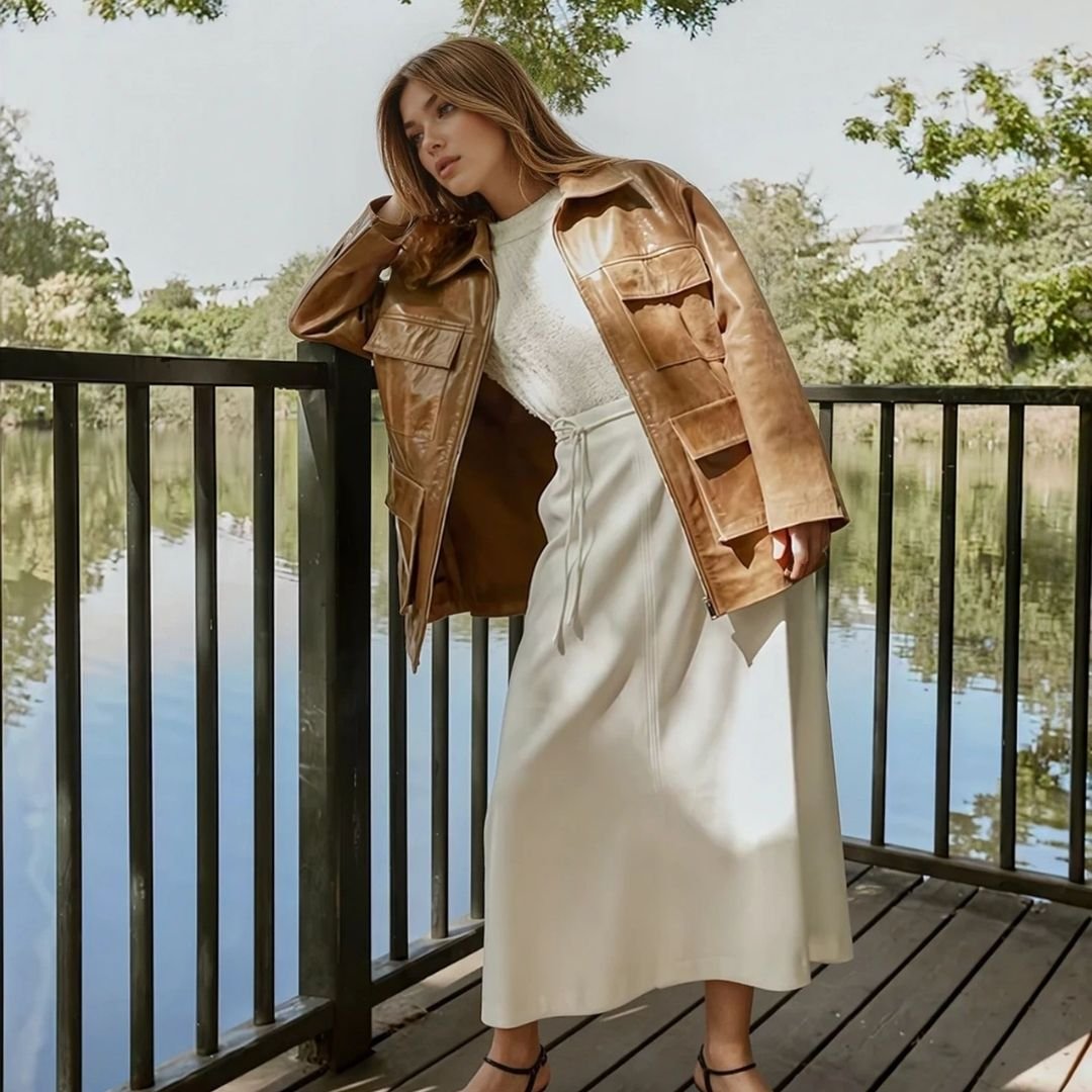 «Γυναίκα με καστανά μαλλιά ποζάρει σε ξύλινη γέφυρα δίπλα σε λίμνη, φορώντας καφέ δερμάτινο τζάκετ, λευκή μπλούζα με υφή δαντέλας και εκρού midi φούστα με κορδόνι στη μέση. Το look ολοκληρώνεται με μαύρα πέδιλα. Το φόντο είναι φυσικό, με δέντρα και γαλήνιο νερό που αντικατοπτρίζει το φως της ημέρας.»