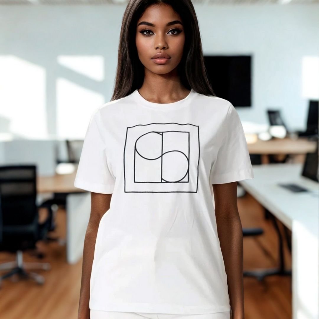 Γυναίκα με μακριά σκούρα μαλλιά φοράει λευκό t-shirt με μίνιμαλ μαύρο γεωμετρικό σχέδιο στο στήθος, σε μοντέρνο επαγγελματικό περιβάλλον γραφείου με φυσικό φωτισμό και ξύλινο πάτωμα. Το στυλ είναι casual-chic, με καθαρή, κομψή αισθητική.