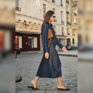 «Γυναίκα περπατά σε αστικό περιβάλλον φορώντας denim πουκαμισοφόρεμα σε dark indigo απόχρωση, συνδυασμένο με μαύρες flat γόβες και καφέ suede τσάντα πλάτης.»