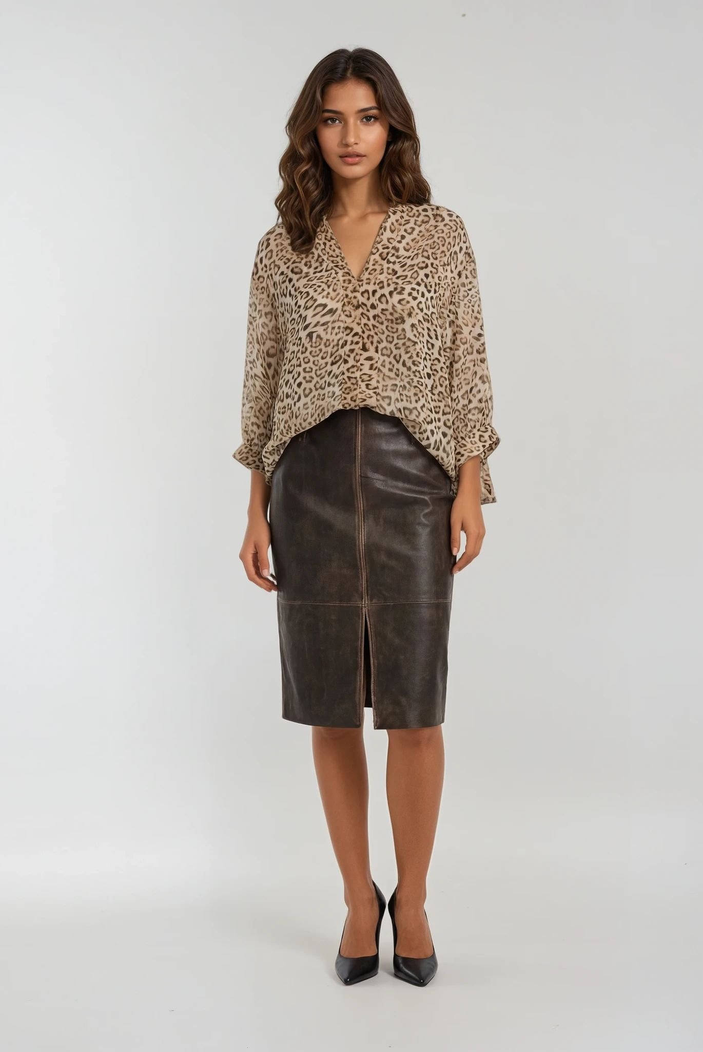 Γυναικείο σύνολο με λεοπάρ μπλούζα σε loose γραμμή και V λαιμόκοψη, συνδυασμένη με καφέ δερματίνη μίντι φούστα σε ίσια γραμμή με μπροστινό σκίσιμο. Το look ολοκληρώνεται με μαύρες γόβες, σε ουδέτερο studio φόντο που αναδεικνύει το κομψό και δυναμικό ύφος.
