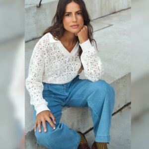 Γυναίκα κάθεται σε τσιμεντένια σκαλοπάτια, φορώντας λευκό πλεκτό μπλουζάκι με διάτρητο μοτίβο πάνω από λευκό πουκάμισο και τζιν παντελόνι σε χαλαρή γραμμή. Έχει μακριά καστανά μαλλιά και κοιτάζει ήρεμα προς την κάμερα, με το χέρι να στηρίζει το πρόσωπό της.