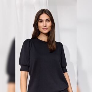 Ημίσωμη φωτογραφία μοντέλου με μαύρη μπλούζα με κοντά φουσκωτά μανίκια ως τον αγκώνα και minimal φόντο με λευκή κουρτίνα.