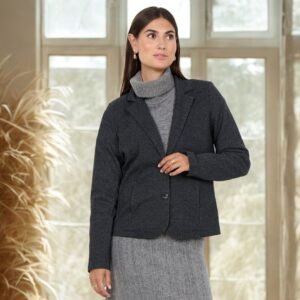 υναίκα φορά ανθρακί blazer με δύο κουμπιά και πέτο, συνδυασμένο με γκρι πλεκτό ζιβάγκο και φούστα στο ίδιο ύφος. Η φωτογράφιση γίνεται σε εσωτερικό, φωτεινό χώρο με μεγάλα παράθυρα και φυσική διακόσμηση από αποξηραμένα φυτά, δημιουργώντας ζεστή, κομψή ατμόσφαιρα.»