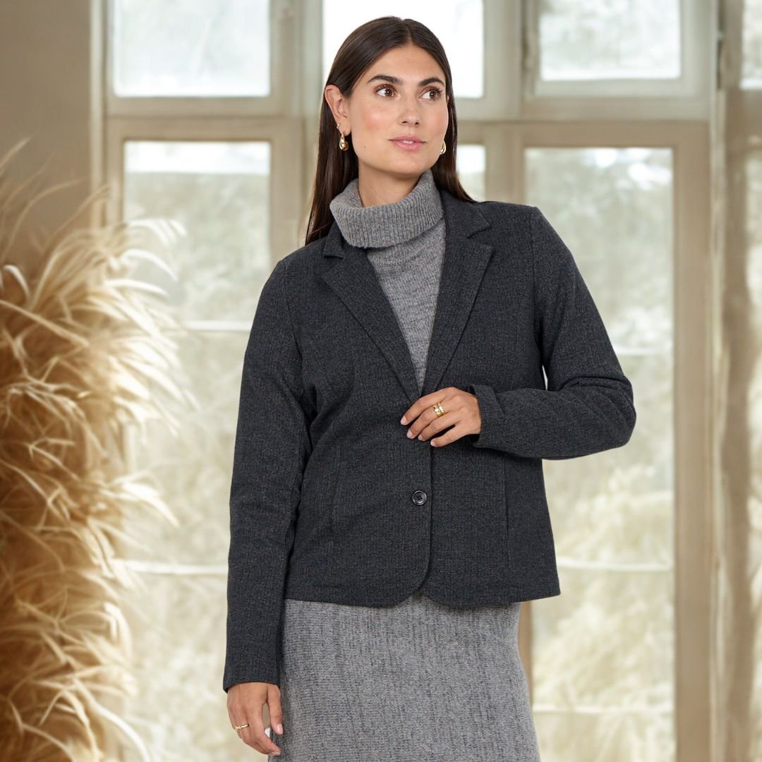 υναίκα φορά ανθρακί blazer με δύο κουμπιά και πέτο, συνδυασμένο με γκρι πλεκτό ζιβάγκο και φούστα στο ίδιο ύφος. Η φωτογράφιση γίνεται σε εσωτερικό, φωτεινό χώρο με μεγάλα παράθυρα και φυσική διακόσμηση από αποξηραμένα φυτά, δημιουργώντας ζεστή, κομψή ατμόσφαιρα.»