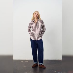 Γυναίκα με ριγέ σοκολατί -εκρού πουκάμισο και τζιν με γυρισμένα ρεβέρ, σε μίνιμαλ στούντιο φόντο, αποπνέοντας casual chic ύφος.