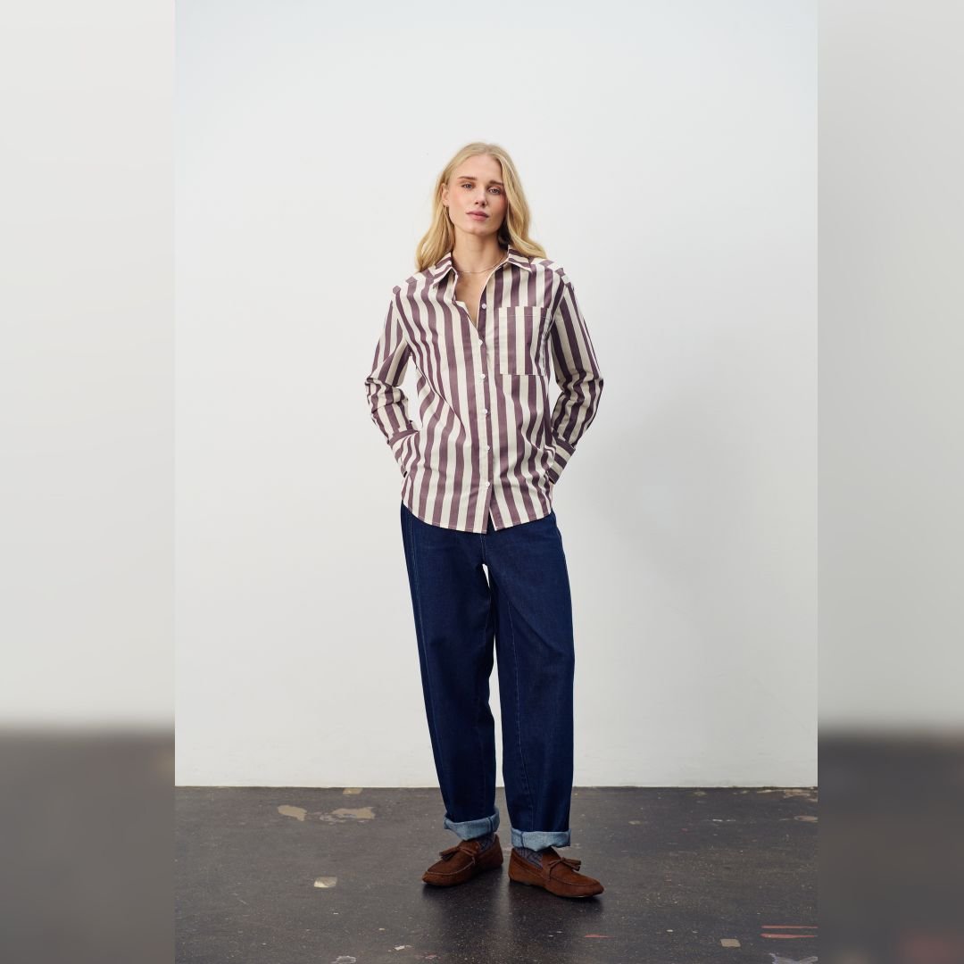 Γυναίκα με ριγέ σοκολατί -εκρού πουκάμισο και τζιν με γυρισμένα ρεβέρ, σε μίνιμαλ στούντιο φόντο, αποπνέοντας casual chic ύφος.