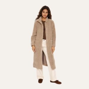 Μακρύ teddy παλτό σε μπεζ απόχρωση φορεμένο ανοιχτό, συνδυασμένο με καφέ τοπ και ανοιχτόχρωμο παντελόνι, σε φωτεινό studio φόντο.