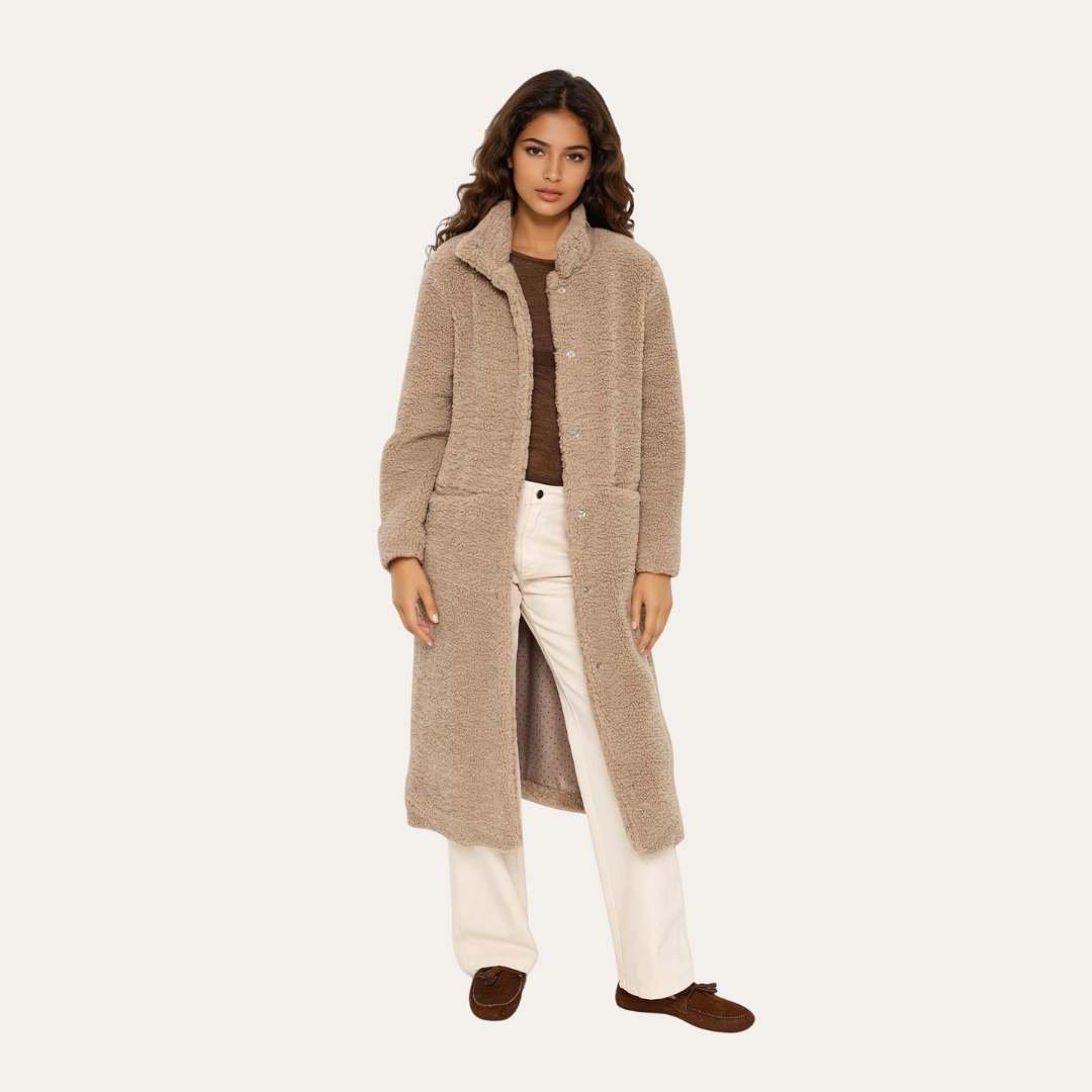 Μακρύ teddy παλτό σε μπεζ απόχρωση φορεμένο ανοιχτό, συνδυασμένο με καφέ τοπ και ανοιχτόχρωμο παντελόνι, σε φωτεινό studio φόντο.