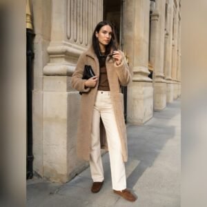 «Γυναίκα περπατά σε αστικό πεζοδρόμιο μπροστά από πέτρινο κτίριο με κολόνες. Φορά μακρύ καμηλό teddy παλτό, καφέ τοπ, εκρού ίσιο παντελόνι και καφέ loafers. Κρατά μαύρη clutch τσάντα και γυαλιά ηλίου στο χέρι. Κομψό, casual chic σύνολο σε γήινους τόνους.»