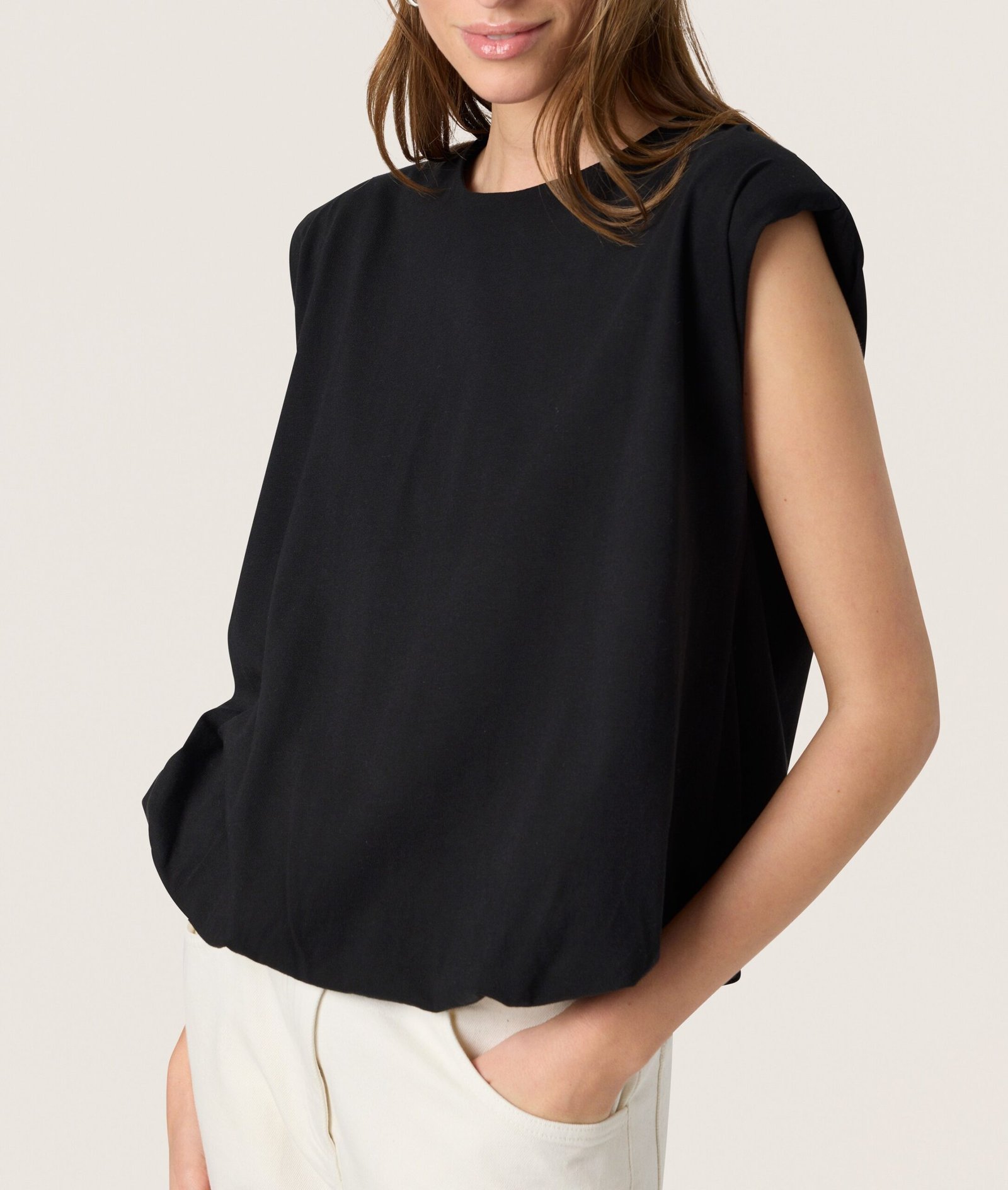 Μοντέλο φορά μαύρη αμάνικη μπλούζα με χαλαρή εφαρμογή, συνδυασμένο με ανοιχτόχρωμο παντελόνι για minimal κομψό καθημερινό styling σε ουδέτερο studio φόντο.