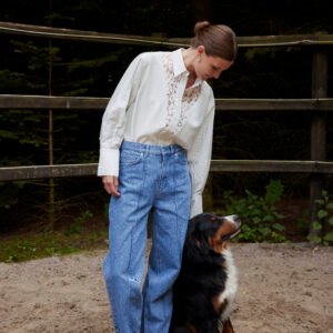 Γυναικείο μπλε denim παντελόνι με μπροστινές ραφές και ψηλόμεση εφαρμογή, συνδυασμένο με εκρού πουκάμισο με δαντελένια λεπτομέρεια, μοντέλο σε φυσικό εξωτερικό χώρο με δέντρα και ξύλινο φράχτη δίπλα σε μεγάλο σκύλο.