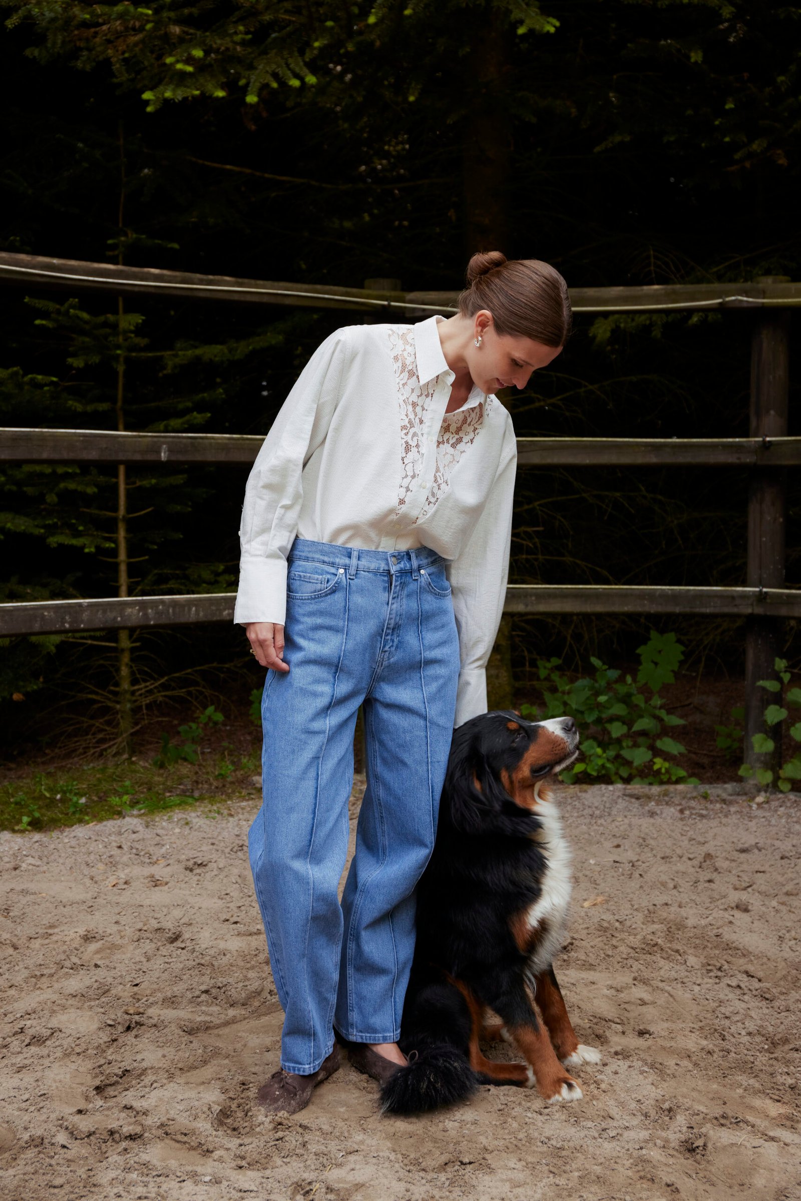 Γυναικείο μπλε denim παντελόνι με μπροστινές ραφές και ψηλόμεση εφαρμογή, συνδυασμένο με εκρού πουκάμισο με δαντελένια λεπτομέρεια, μοντέλο σε φυσικό εξωτερικό χώρο με δέντρα και ξύλινο φράχτη δίπλα σε μεγάλο σκύλο.