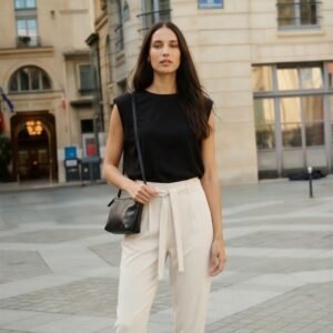 Alt text Γυναίκα φορά εκρού ψηλόμεσο παντελόνι με ζώνη και μαύρο αμάνικο top, συνδυασμένο με μαύρη τσάντα ώμου, σε κομψό urban look, φωτογραφημένη σε ευρωπαϊκό δρόμο πόλης με κλασικά κτίρια στο φόντο.