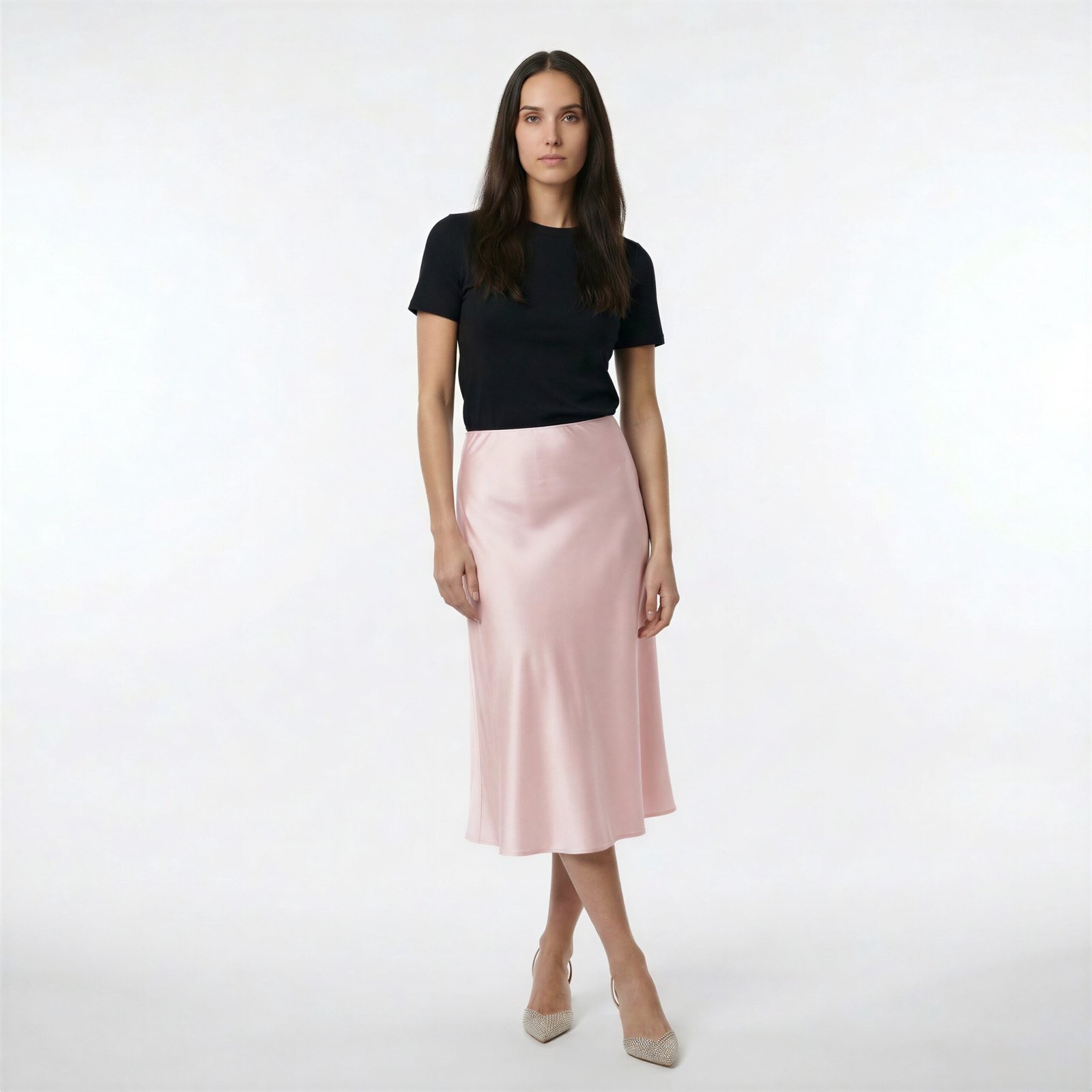 Γυναίκα φορά μίντι σατέν φούστα σε ροζ πούδρα με απαλή, ρευστή υφή και ελαφρώς Α-line εφαρμογή, συνδυασμένη με μαύρο fitted t-shirt και μυτερές γόβες, σε καθαρό λευκό studio φόντο.