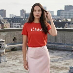 Γυναίκα σε ταράτσα πόλης φορά μίντι σατέν φούστα σε ροζ πούδρα με απαλή υφή και ελαφρώς Α-line εφαρμογή, συνδυασμένη με κόκκινο t-shirt, σε urban minimal φόντο με κτίρια στο βάθος.