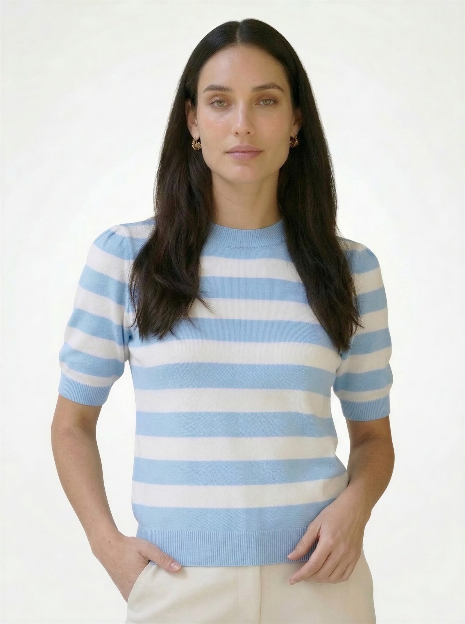 Γυναικείο ριγέ κοντομάνικο πλεκτό top σε απαλό γαλάζιο και λευκό με στρογγυλή λαιμόκοψη και rib τελείωμα, εφαρμοστή γραμμή, φορεμένο σε μοντέλο με μπεζ παντελόνι σε φωτεινό studio φόντο.