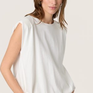 Γυναίκα φορά λευκή oversized μπλούζα με βάτες στους ώμους και balloon εφαρμογή, συνδυασμένη με λευκή βερμούδα σε minimal studio με ουδέτερο φόντο.