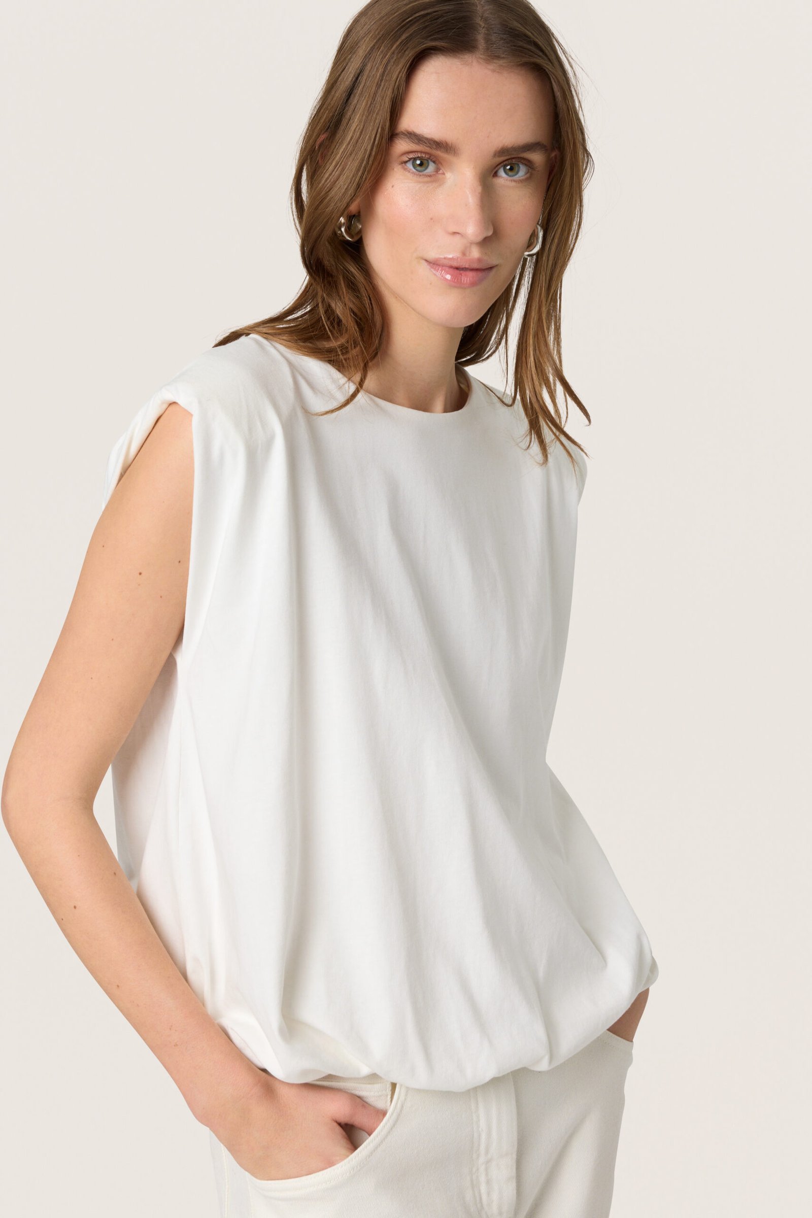 Γυναίκα φορά λευκή oversized μπλούζα με βάτες στους ώμους και balloon εφαρμογή, συνδυασμένη με λευκή βερμούδα σε minimal studio με ουδέτερο φόντο.