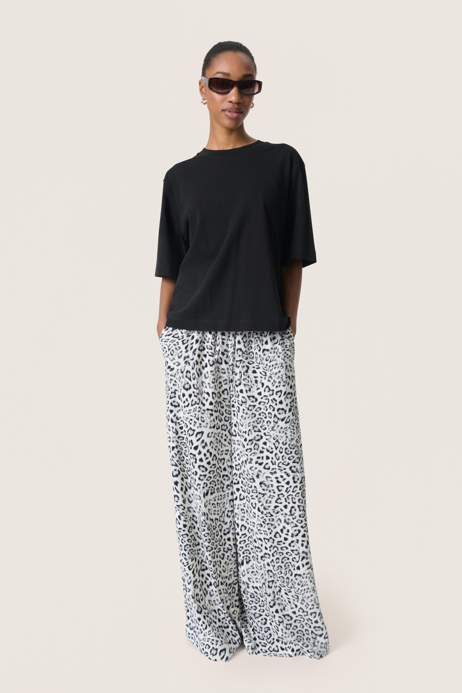 Wide leg παντελόνα με ασπρόμαυρο leopard print συνδυασμένη με μαύρο T-shirt και μοντέρνα γυαλιά ηλίου. Casual chic εμφάνιση σε μπεζ studio φόντο.